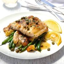 Fish Piccata with smashed potatoes and steamed greens on a white plate with lemon wedges, ready to be eaten.